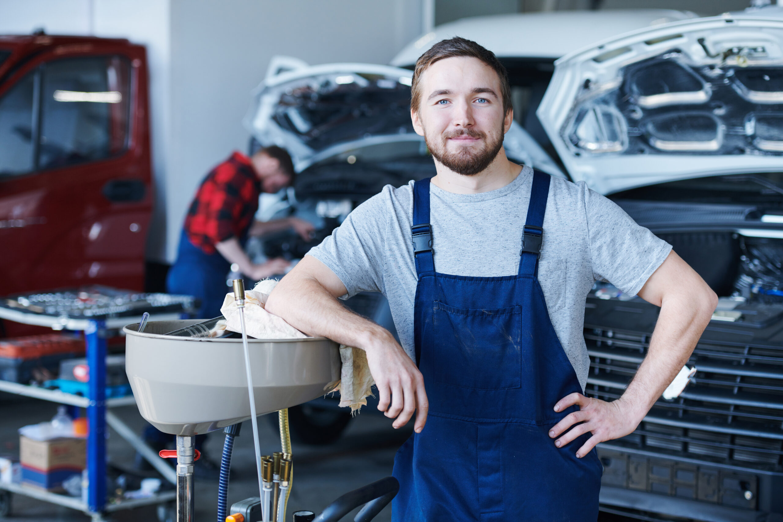 Happy young repairman in blue overalls looking at you in workshop on background of working colleague